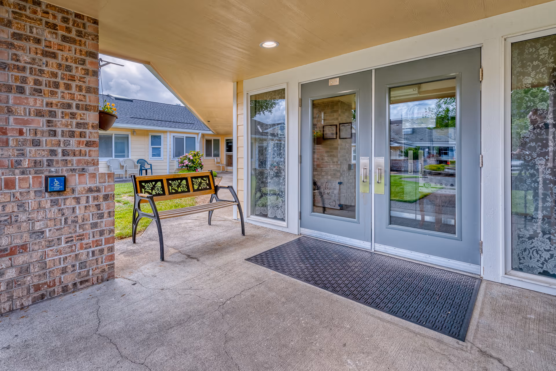 Covered entrance area of a senior living facility with double glass doors, a decorative bench, a brick wall with a handicap accessibility sign, and a view of the courtyard with chairs and flowers.