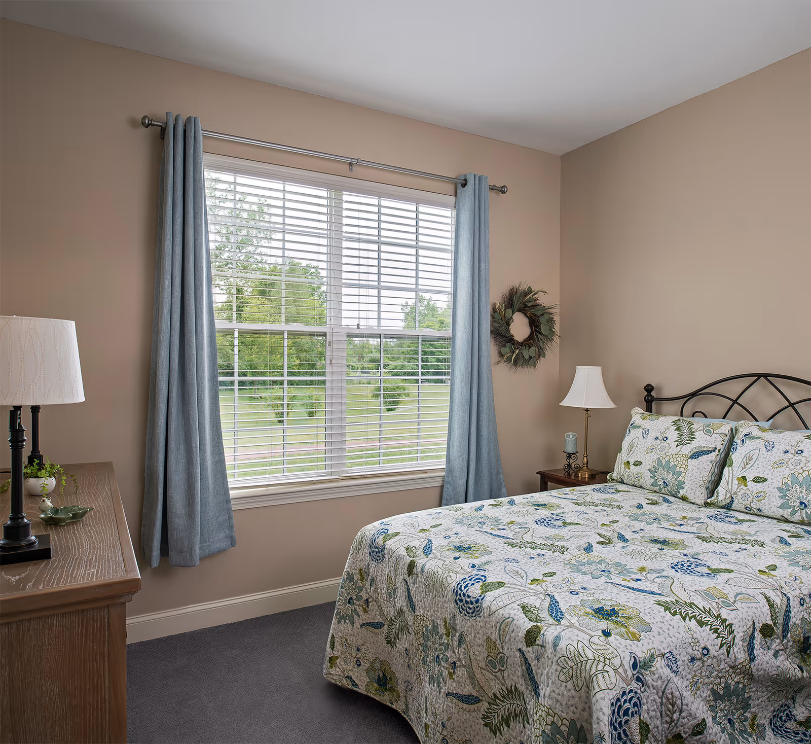 A bedroom with a large window covered by blue curtains and white blinds. The bed has a floral patterned bedspread and matching pillows. There is a wooden dresser with a lamp and small plant on it, and a nightstand with a lamp and candle. A decorative wreath hangs on the beige wall.