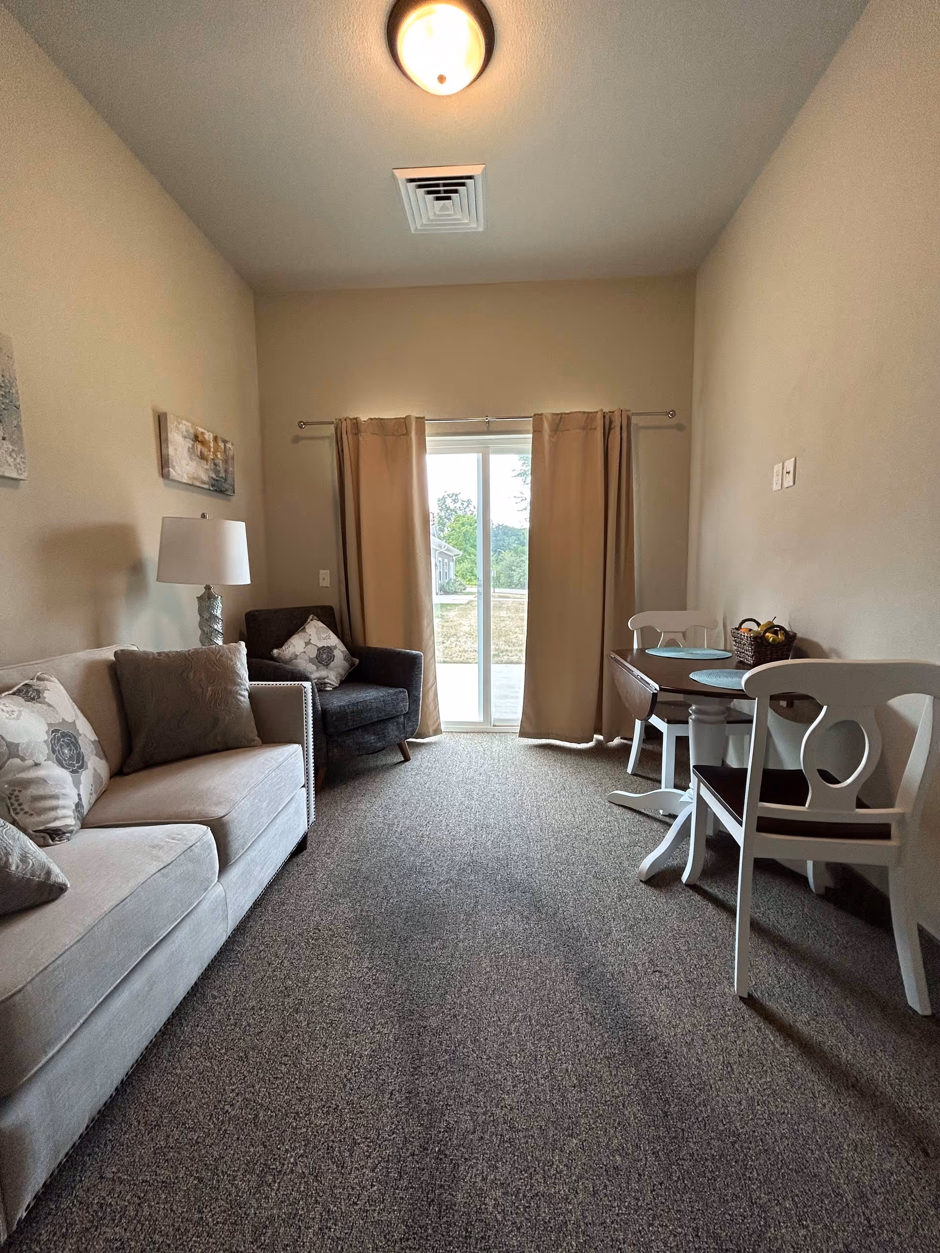 A small living area with a beige sofa and patterned pillows on the left, a dark armchair with a decorative pillow next to a lamp, and a small wooden dining table with two white chairs on the right. Beige curtains cover a sliding glass door that leads outside. The room has beige walls and carpeted floor.