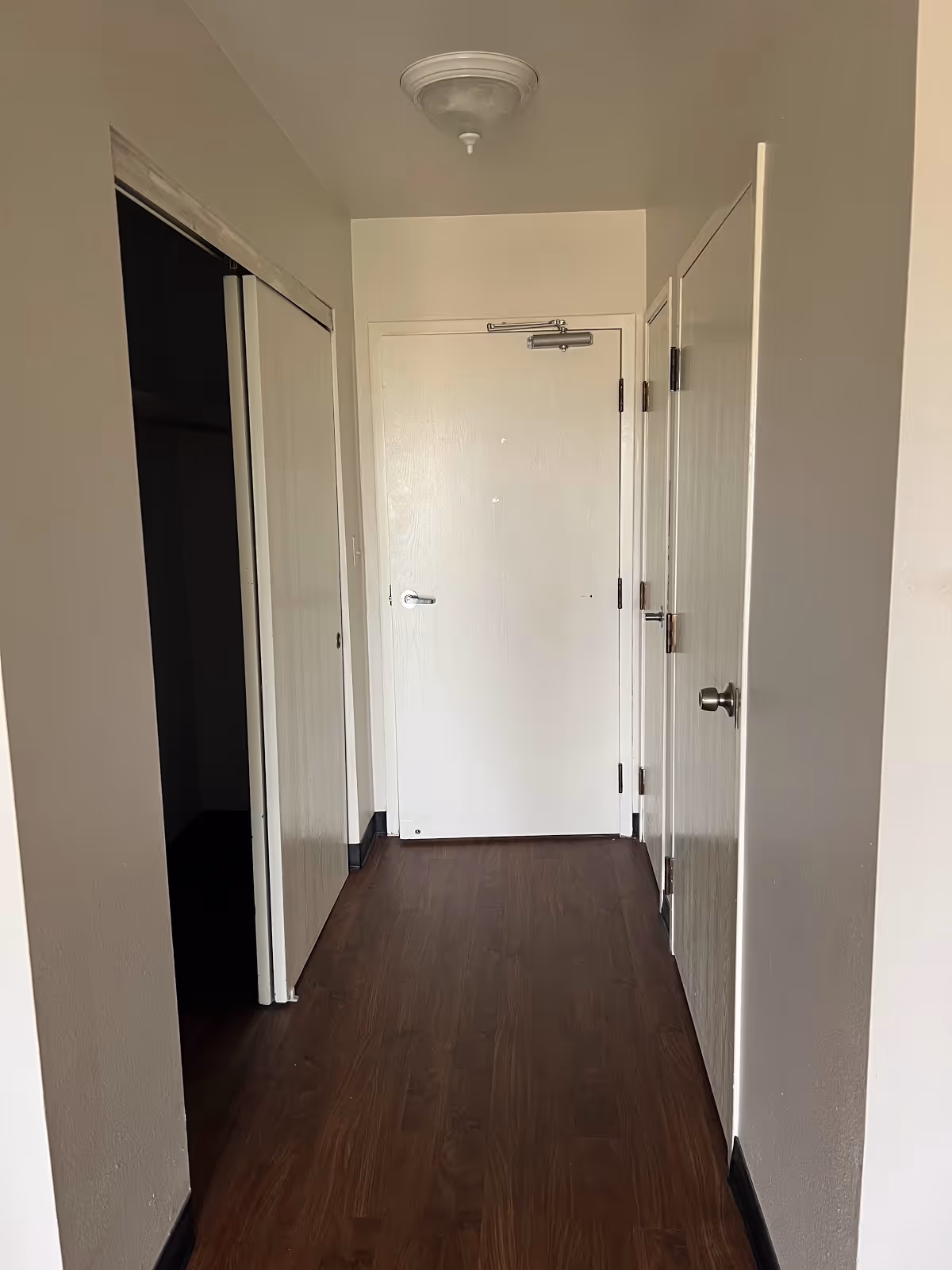A narrow hallway with dark wood flooring and white walls. There are three closed white doors visible: one at the end of the hallway, one on the right side, and one closet door on the left side that is partially open. A ceiling light fixture is mounted above.