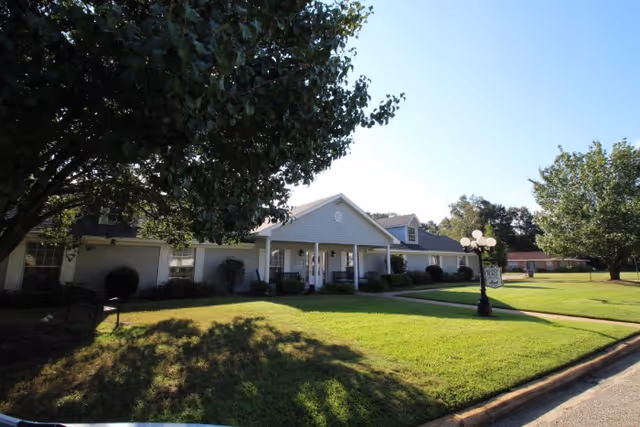 Single-story building with a white exterior and a covered entrance, surrounded by well-maintained green lawns and trees under a clear blue sky.