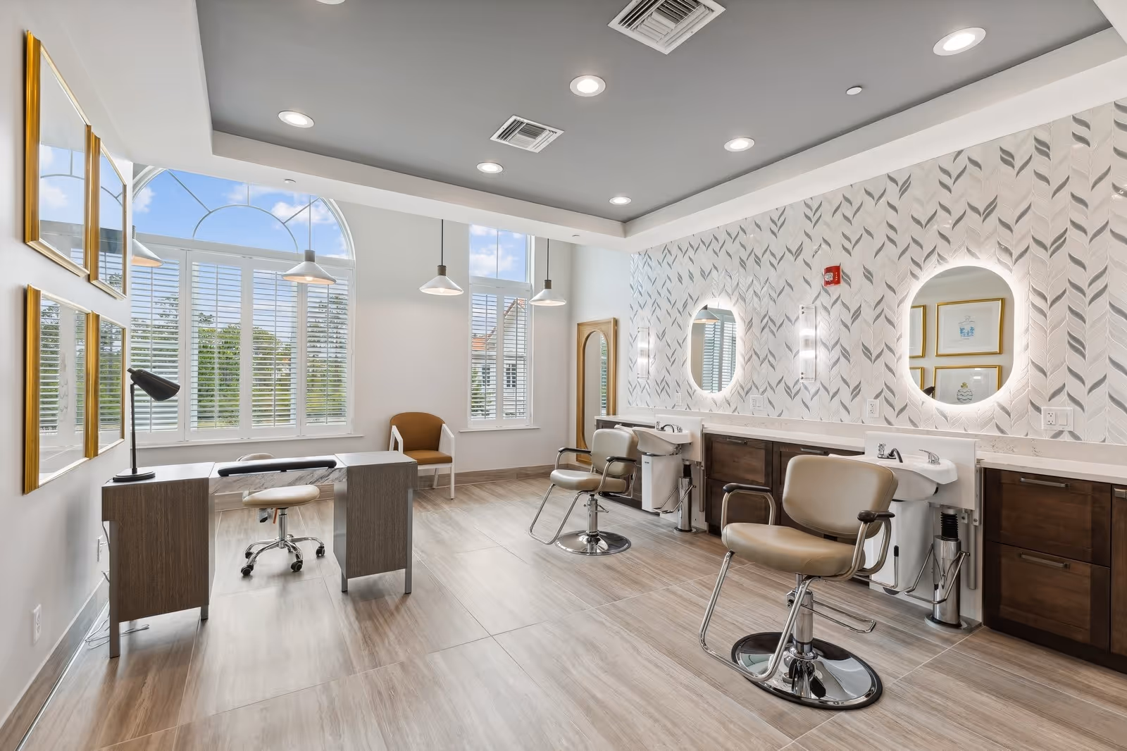 A bright and modern salon area with two styling chairs in front of illuminated oval mirrors mounted on a patterned wall. There are sinks for hair washing beneath the mirrors. The room features large windows with white shutters, a desk with a rolling stool, and a single armchair near the windows. The ceiling has recessed lighting and pendant lights hanging above the desk.