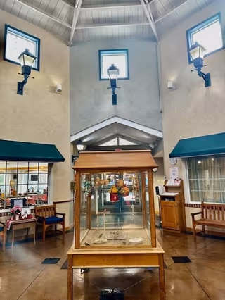 Interior view of a senior living facility lobby or common area with high ceilings and large windows near the roof. There is a wooden display case with decorative items in the center, flanked by wooden benches and small tables with chairs. Wall-mounted lantern-style lights and green awnings over windows add to the decor.