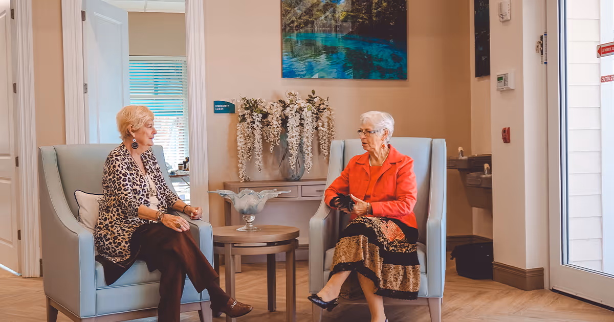 Two elderly women sitting and conversing in a well-lit sitting area with light blue armchairs, a small round table between them, a decorative vase with white flowers on a console table behind them, and a large nature-themed painting on the wall.