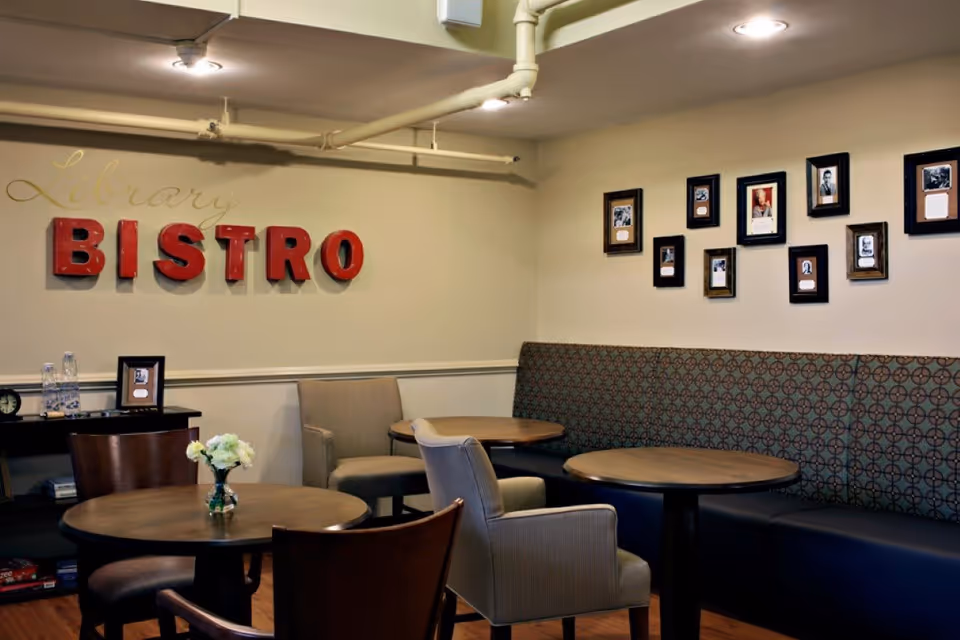 Interior view of a cozy bistro area in a senior living facility with round wooden tables, cushioned chairs, and a patterned bench along the wall. The wall features framed photos and the words 'Library BISTRO' in decorative lettering.