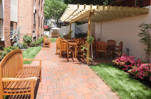 Outdoor patio area at Lester Senior Living with wooden benches and tables under a pergola, surrounded by brick walls, green grass, and flowering plants.