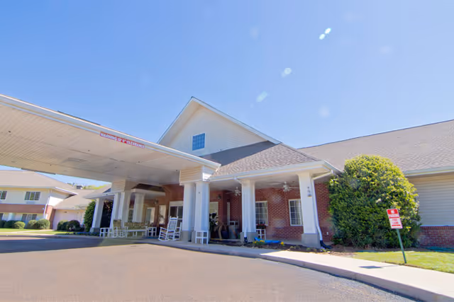 Exterior view of a senior living facility with a covered entrance supported by white columns. The building has a brick facade with white trim and a sloped roof. There are rocking chairs on the porch and a small green bush near the entrance. A sign indicating parking rules is visible on the lawn.