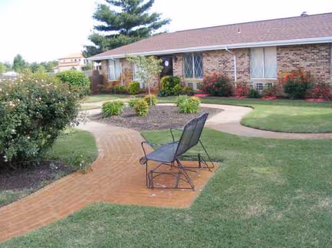 Brick building exterior with a curved brick path, garden beds, and a metal chair on patio.