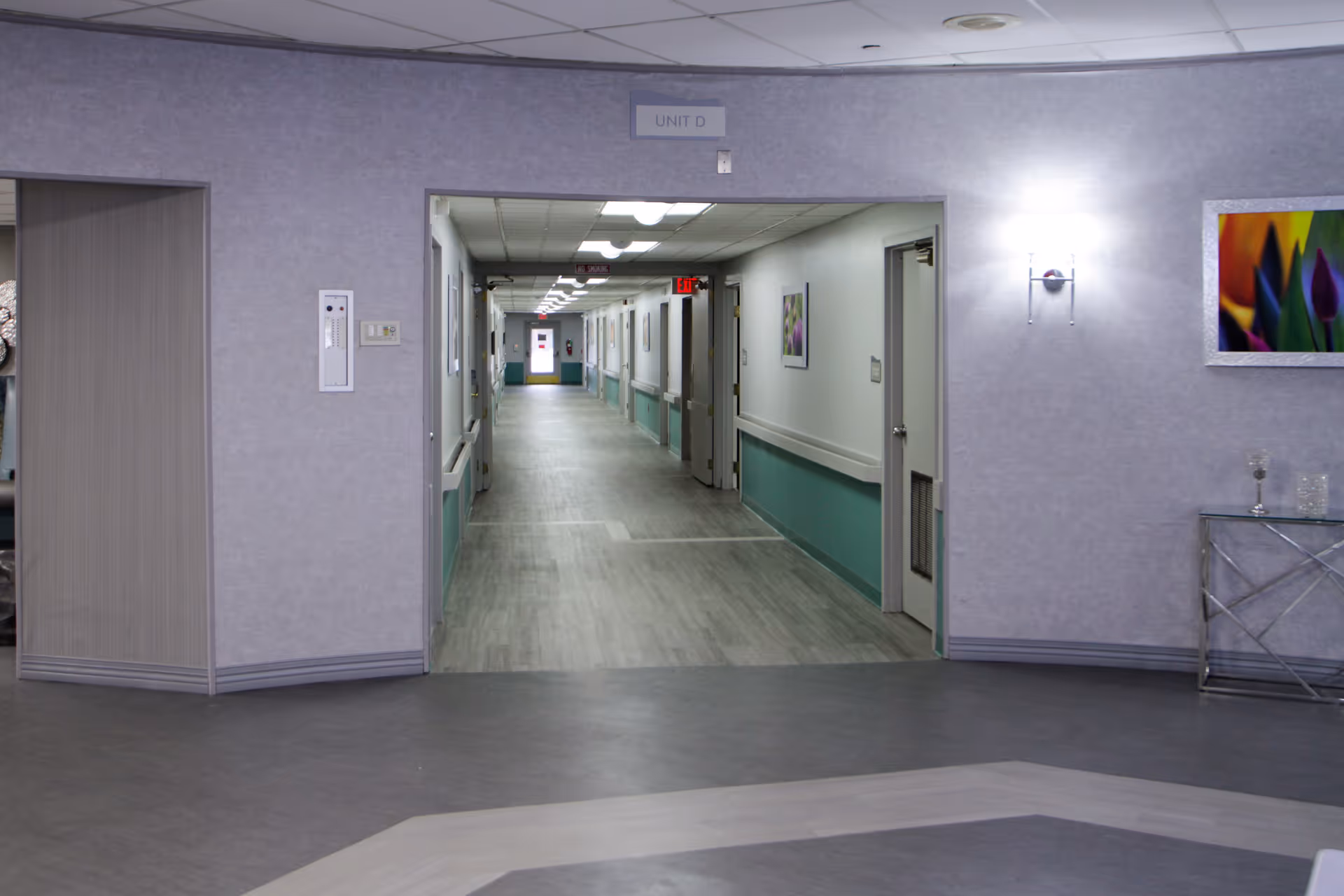 A clean, well-lit hallway in a senior living facility with light gray and teal walls, handrails along the sides, and several doors leading to rooms. The hallway is viewed from a larger room with purple walls, a framed colorful flower picture, a wall sconce light, and a small glass table with decorative items. A sign above the hallway entrance reads 'UNIT D'.
