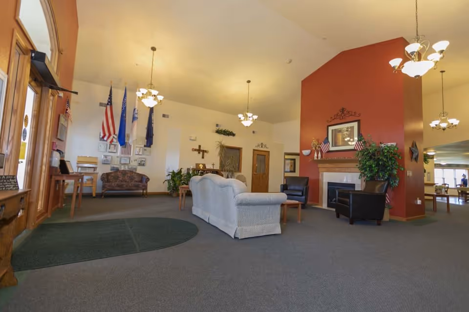 Spacious assisted living facility common area with a high ceiling, carpeted floor, and multiple seating arrangements including a light gray sofa and black armchairs around a fireplace with a red accent wall. American flags and other flags are displayed on the far wall above a wooden bench and framed pictures. Several hanging light fixtures illuminate the room, and there are plants and decorative items placed throughout the space.