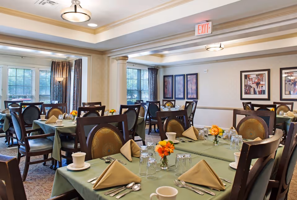 A dining room in a senior living facility with multiple tables covered in green tablecloths, each set with beige folded napkins, cups, glasses, and silverware. The room has large windows with curtains, framed artwork on the walls, and ceiling lights providing warm illumination.