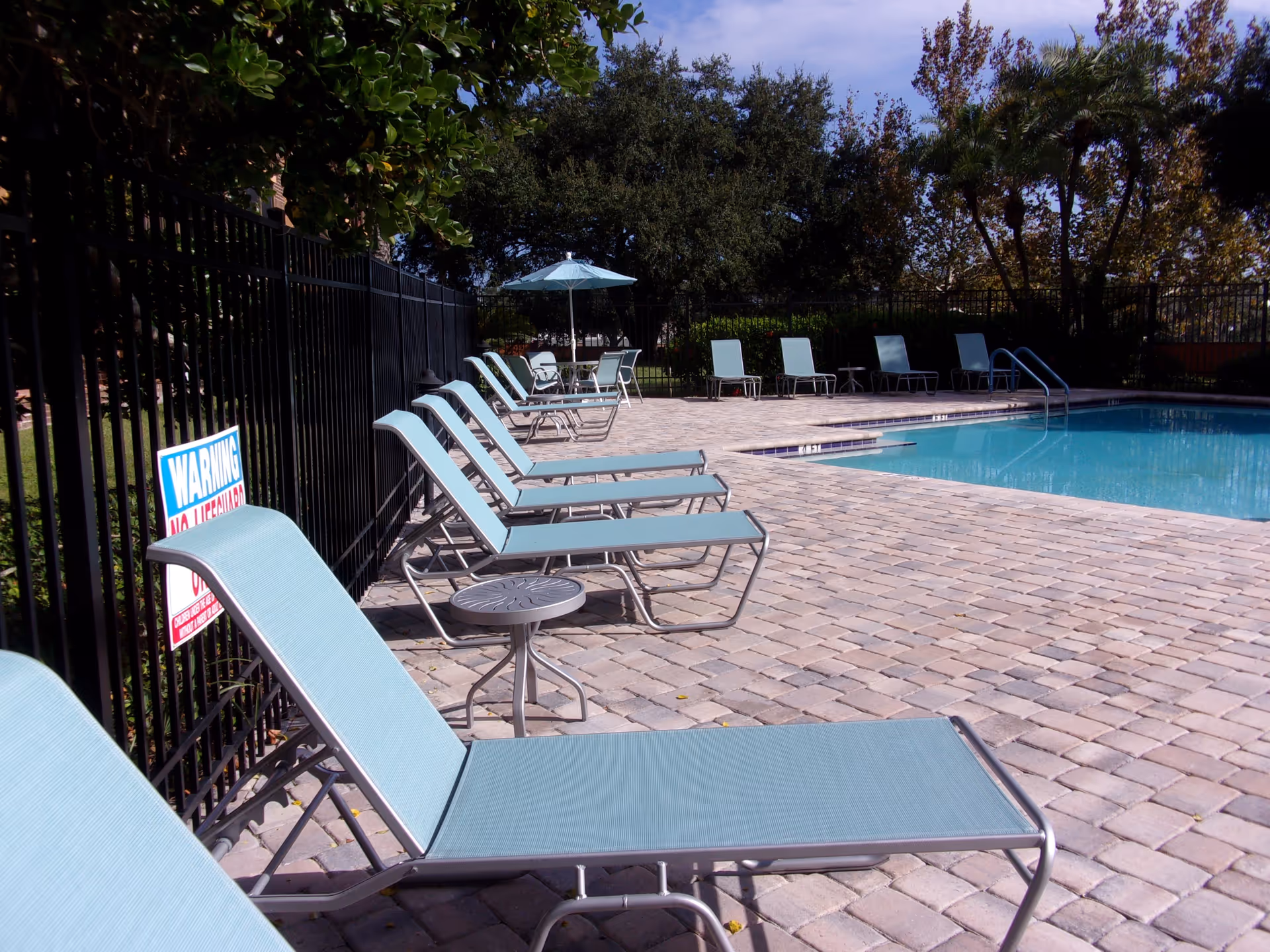 Outdoor swimming pool area with several light blue lounge chairs lined up on a paved pool deck. There is a small round table between some of the chairs and a blue umbrella shading a table with chairs in the background. Trees and greenery surround the fenced pool area under a clear sky.