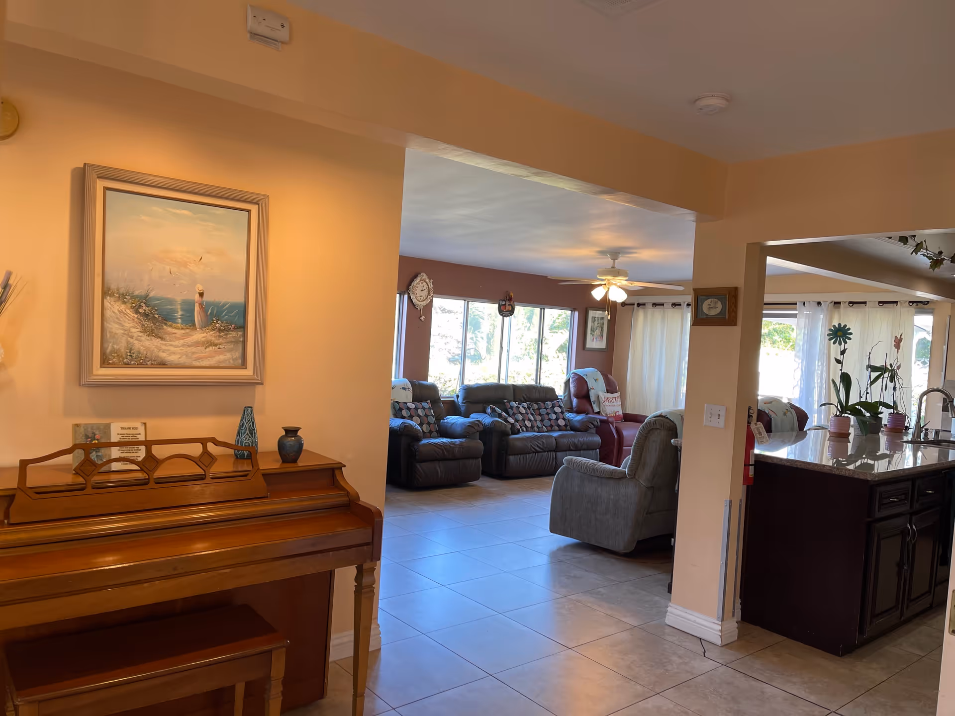 Interior view of a senior living facility showing a wooden piano with a bench and a framed painting above it on the left. The room opens into a living area with several recliner chairs arranged near large windows with curtains. To the right, there is a kitchen area with a granite countertop, sink, and potted plants. The floor is tiled and the walls are painted in warm tones.