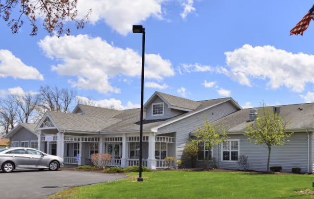 Exterior view of a single-story senior living facility building with gray siding, white trim, and a gabled roof under a partly cloudy blue sky. There is a parking area with a silver car, a black lamp post in front, and a well-maintained green lawn with small trees and shrubs.