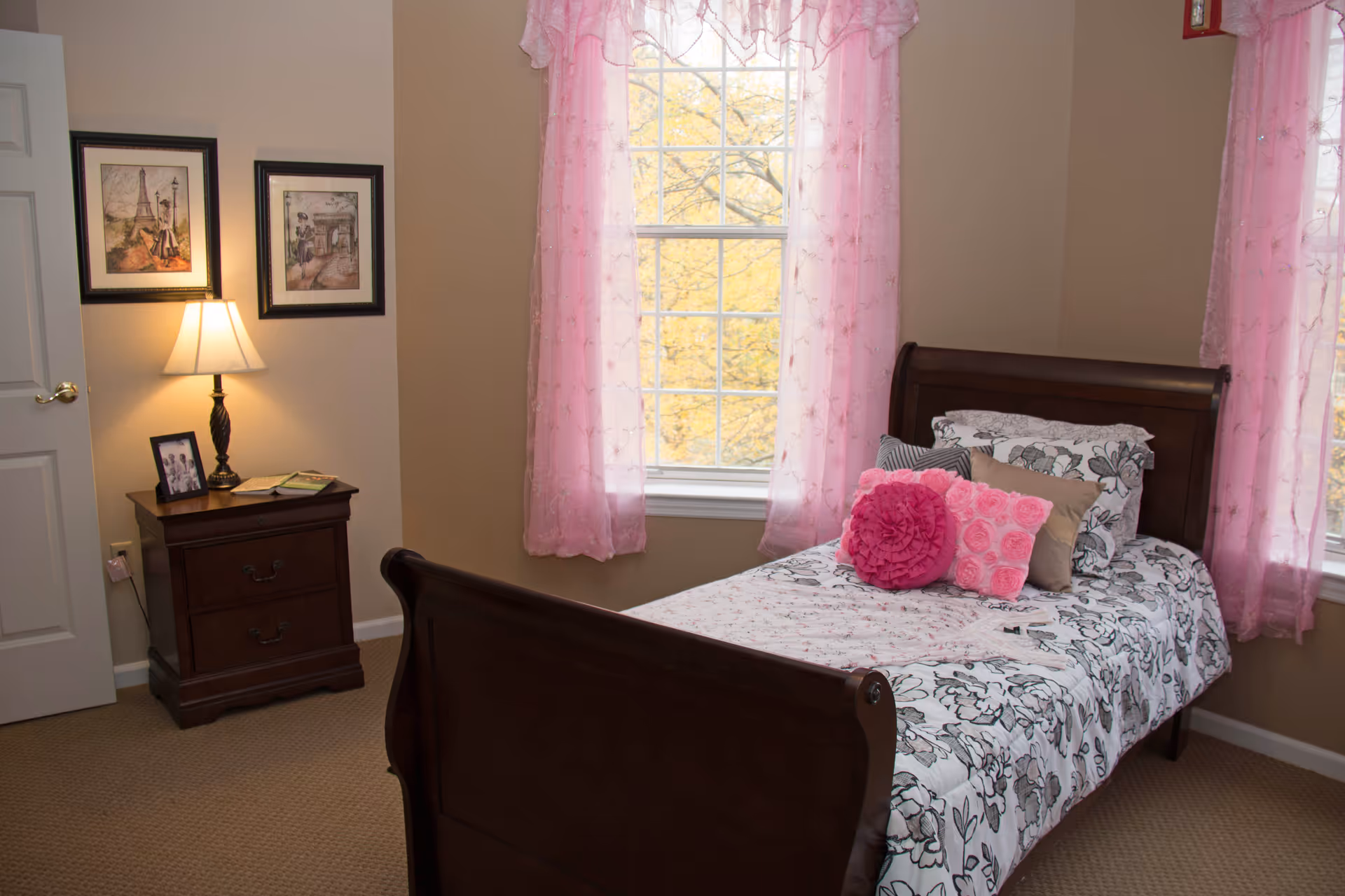 A cozy bedroom with a dark wooden bed frame and a floral patterned bedspread. The bed is decorated with several pillows in shades of pink and beige. There are two windows with sheer pink curtains allowing natural light to enter. Next to the bed is a dark wooden nightstand with a lamp, a framed photo, and some books. Two framed pictures hang on the wall above the nightstand.
