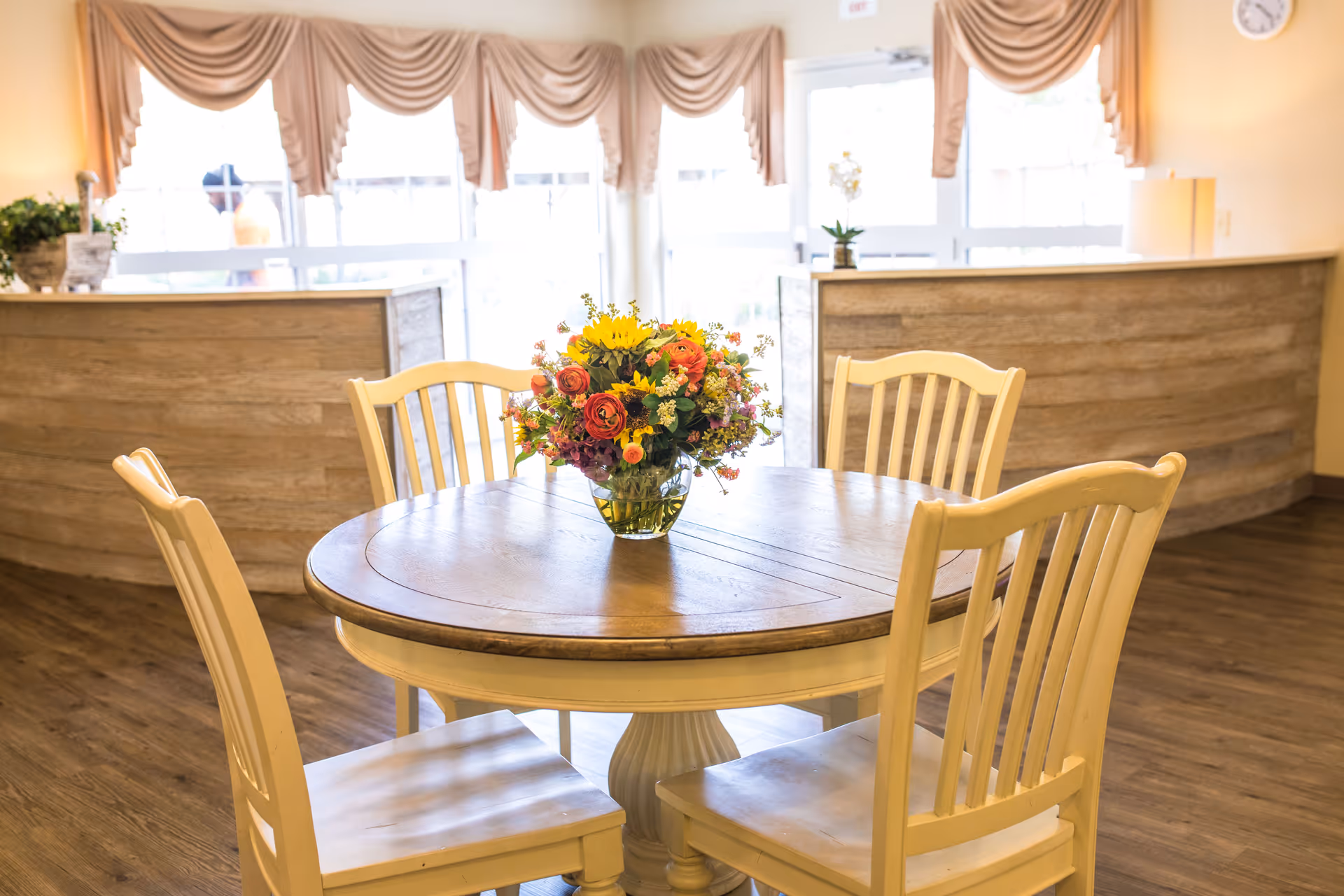 A round wooden table with a vase of colorful flowers in the center, surrounded by four cream-colored wooden chairs. The background shows a reception desk with wood paneling, large windows with beige curtains, and a door leading outside.