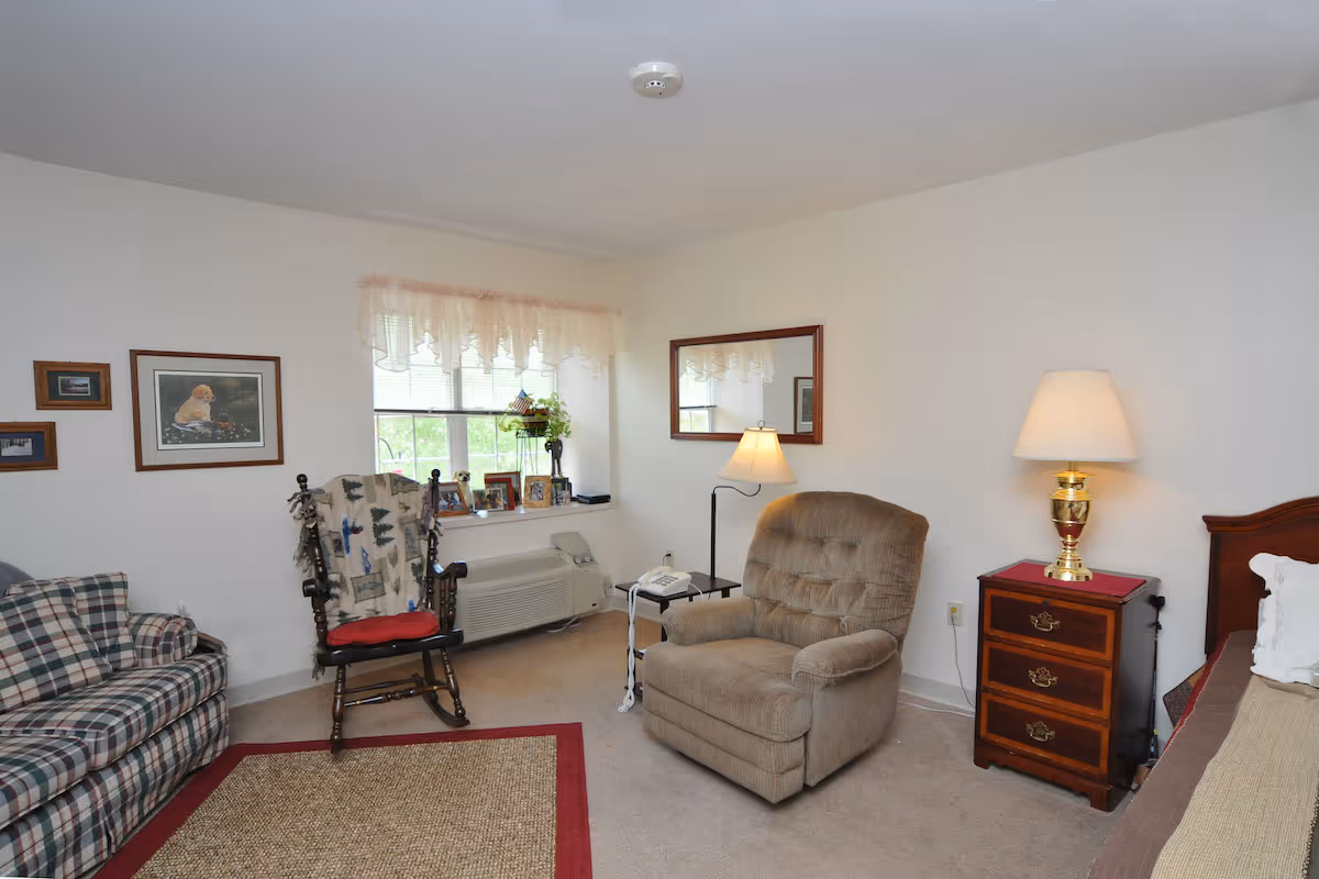 A cozy living area with a plaid sofa, a wooden rocking chair with a blanket, a cushioned armchair, a wooden nightstand with a lamp, and a bed partially visible on the right. There is a window with sheer curtains, framed pictures on the wall, and a mirror above the armchair.