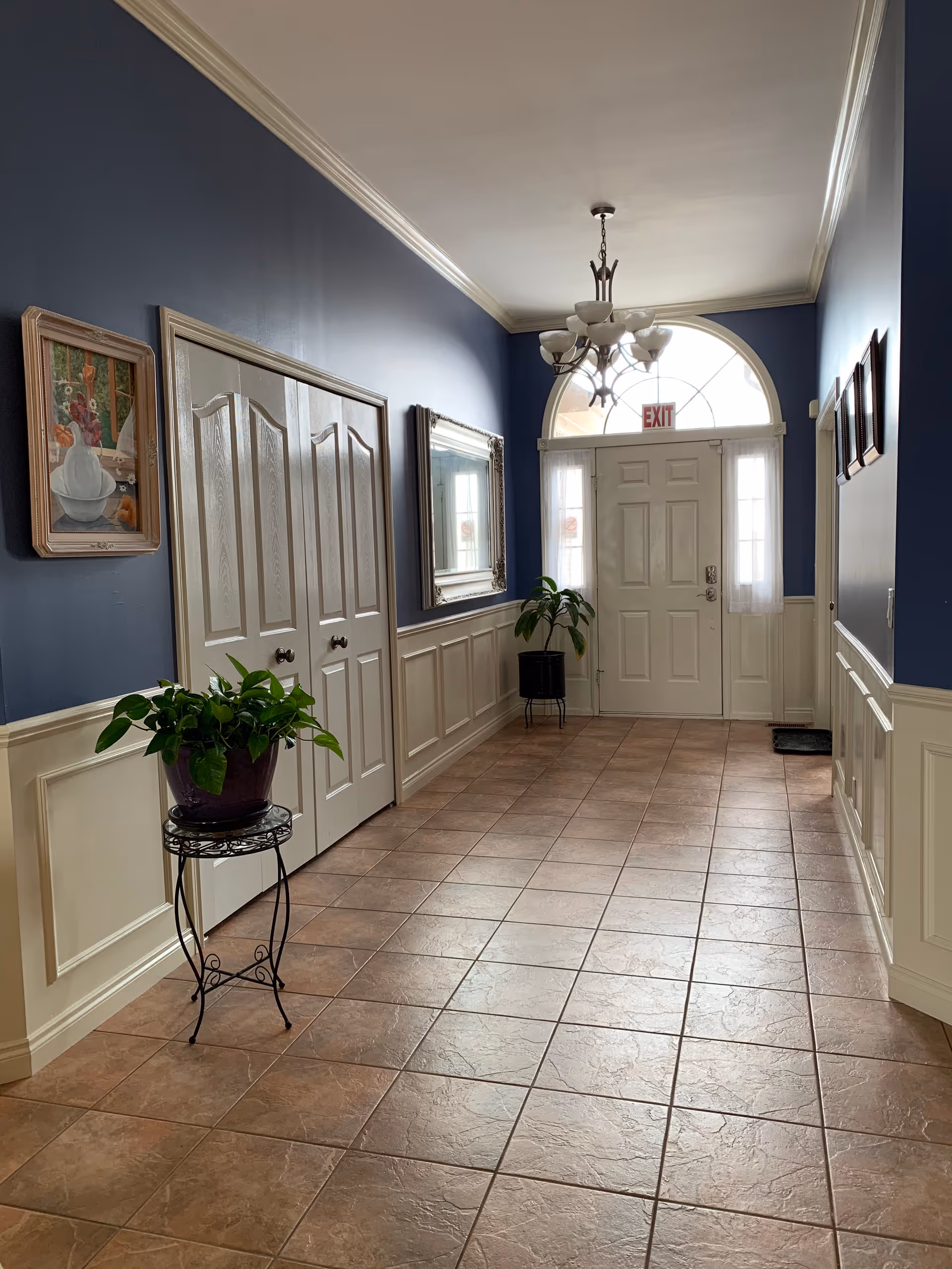 Bright tiled entry hallway with blue walls, white wainscoting, potted plants, chandelier, and a front door with an EXIT sign.