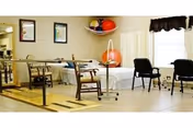 A rehabilitation room with parallel bars for walking exercises, chairs, a hospital bed, and exercise balls on a shelf. The room has tiled floors, framed pictures on the wall, and a window with a curtain.