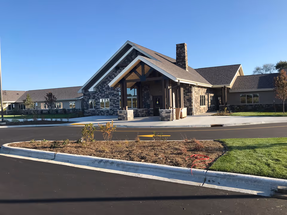 Exterior view of a single-story senior living facility building with stone and siding facade, a covered entrance with wooden beams, a chimney, and a paved driveway with landscaped areas and grass under a clear blue sky.