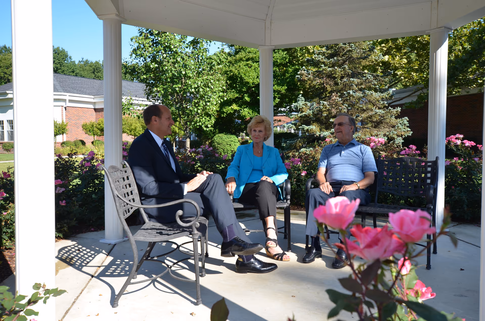 Three people sitting and conversing under a white gazebo in a garden area with blooming pink flowers and green trees in the background at Woodhaven Retirement Community.