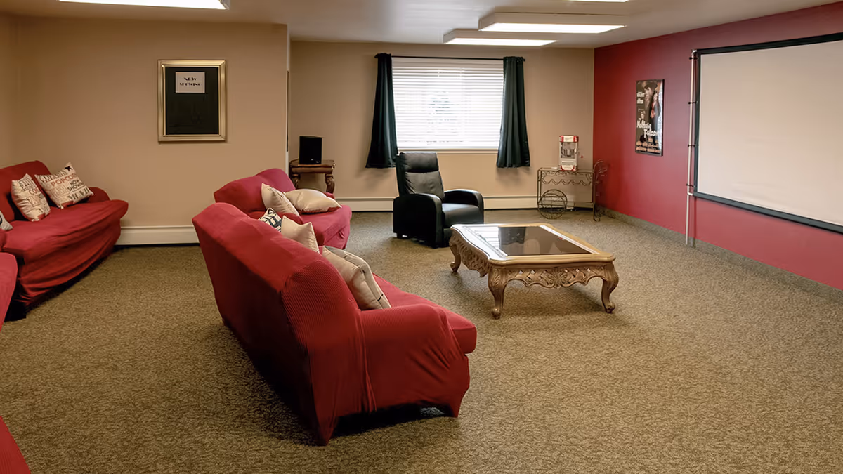A cozy living room with red couches covered in slipcovers, a black recliner chair, a wooden coffee table with a glass top, and a large projector screen on a red accent wall. There is a window with dark curtains, a popcorn machine on a metal stand, and a framed sign on the beige wall.