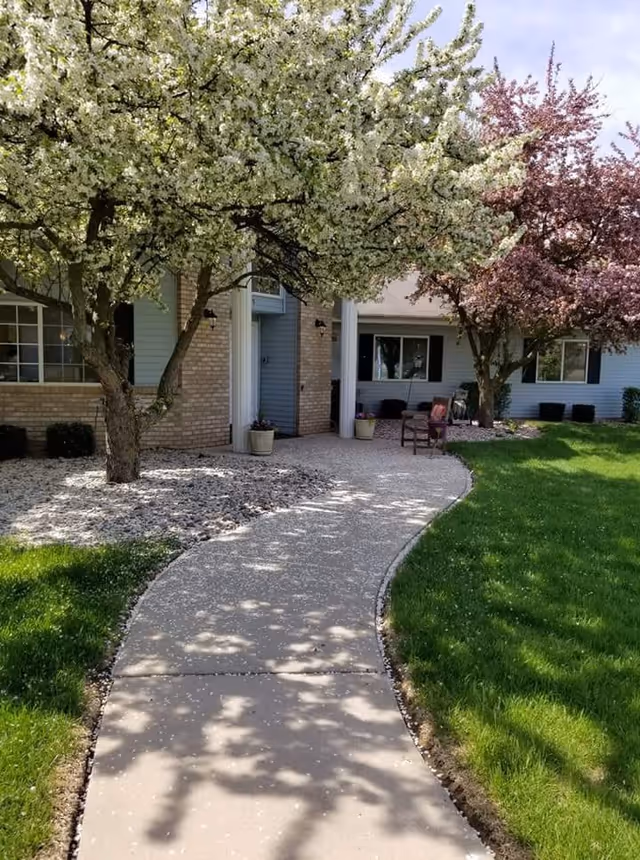 A curved concrete walkway leading to the entrance of a building with light blue siding and brick accents. The walkway is bordered by green grass and landscaped areas with white rocks. Two trees with blooming white and pink flowers provide shade along the path. There are potted plants near the entrance and outdoor chairs on a small patio area.