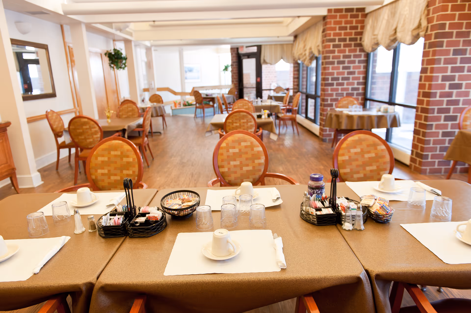 A dining room in a senior living facility with tables covered in brown tablecloths, set with white cups, glasses, napkins, and condiment baskets. The room has wooden floors, patterned chairs, large windows with valances, and brick accent walls.