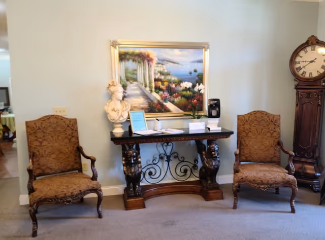 A sitting area with two ornate upholstered armchairs on either side of a decorative wooden console table. On the table are a bust sculpture, a framed sign, some papers, a small plant, and a coffee maker. Above the table hangs a large framed painting of a scenic garden pathway overlooking the sea. To the right of the chairs is a tall wooden grandfather clock.