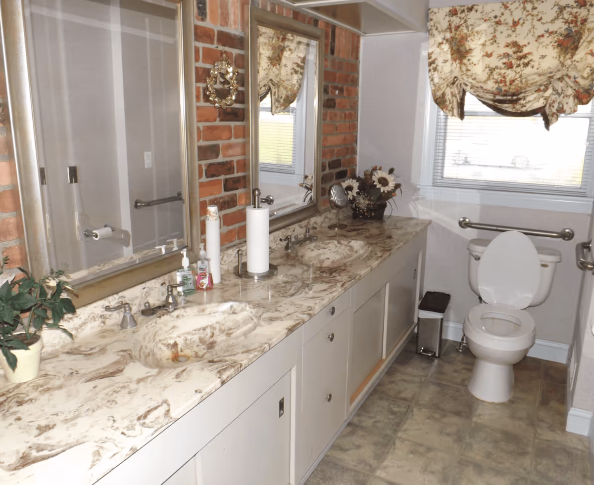 A bathroom with a long marble countertop featuring two sinks, two large mirrors above the sinks, and various items including hand soap, paper towels, and a small plant. The wall behind the sinks has a brick accent. There is a toilet with grab bars on both sides near a window with floral curtains. The floor is tiled.