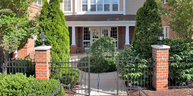 Gated front entrance of a brick senior living building with brick pillars, iron fence, landscaped walkway and a covered porch.