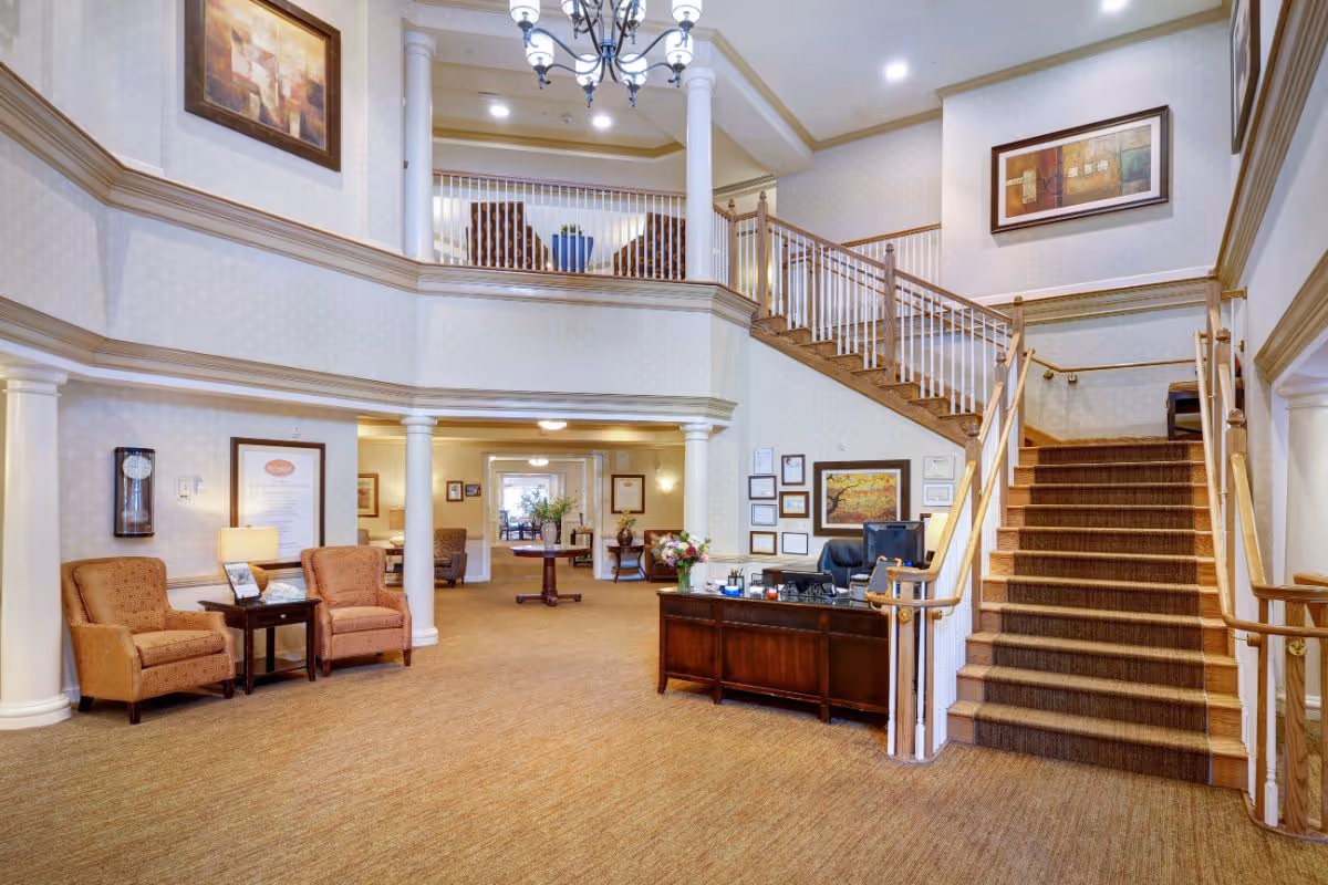 Spacious and well-lit interior of a senior living facility featuring a large staircase with wooden handrails and carpeted steps, a reception desk with a computer, two upholstered chairs with a small table between them, decorative columns, framed artwork on the walls, and a second-floor balcony with additional seating.