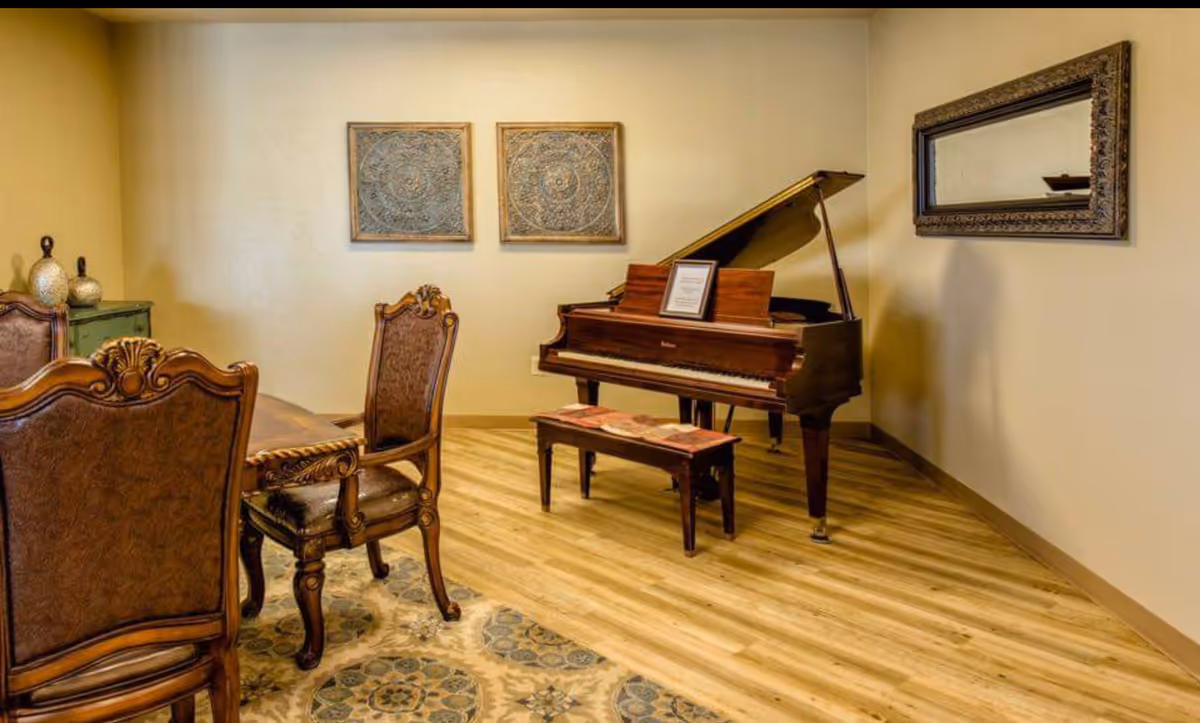 A room with a wooden grand piano and matching bench on a wooden floor. To the left, there is a dining table with ornate wooden chairs. The walls are beige with two decorative framed artworks and a large framed mirror. A patterned rug is partially visible under the dining table.