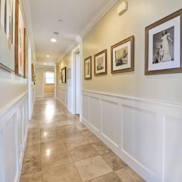 Long bright hallway with tiled floors, white wainscoting and framed artwork along the walls.