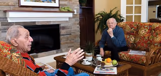 Two elderly men sitting in a cozy living room with patterned armchairs and a stone fireplace. A coffee table in front of them holds a plate of fruit, cups, and a newspaper. One man is gesturing while talking, and the other is listening attentively.