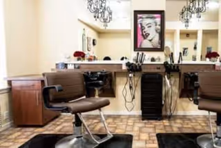 Interior view of a salon area with two brown salon chairs facing mirrors mounted on a beige wall. The space includes hair styling tools such as hair dryers and brushes placed on a counter and a black rolling cart. The floor has a patterned tile design, and two chandeliers hang from the ceiling. A framed picture of Marilyn Monroe is visible on the wall between the mirrors.