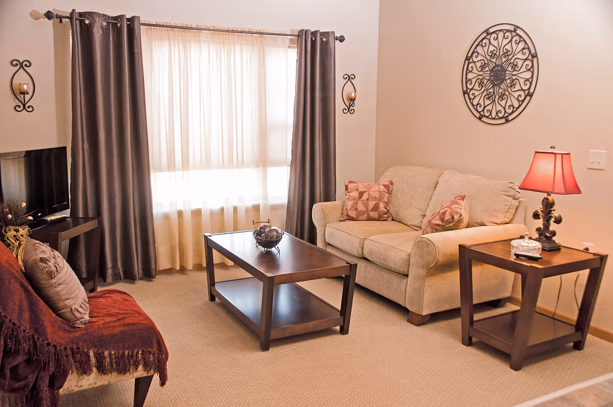 Bright living room with a beige loveseat, dark wood coffee and side tables, a red-shaded lamp, TV, and a window with curtains.