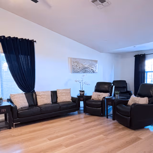 A living room with a black leather sofa and three matching black leather recliners, each with beige decorative pillows. The room has light wood flooring, white walls, two windows with dark blue curtains, and a piece of artwork hanging on the wall above the sofa. There are small side tables with plants and a white orchid in the center.