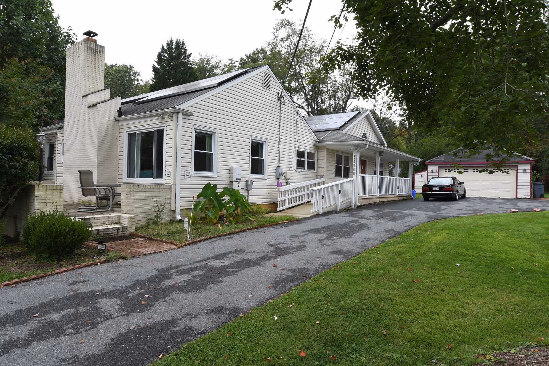 Exterior view of a single-story white house with a chimney, a wheelchair accessible ramp leading to the front porch, a driveway with a parked car, and a detached garage. The house is surrounded by trees and a grassy lawn.