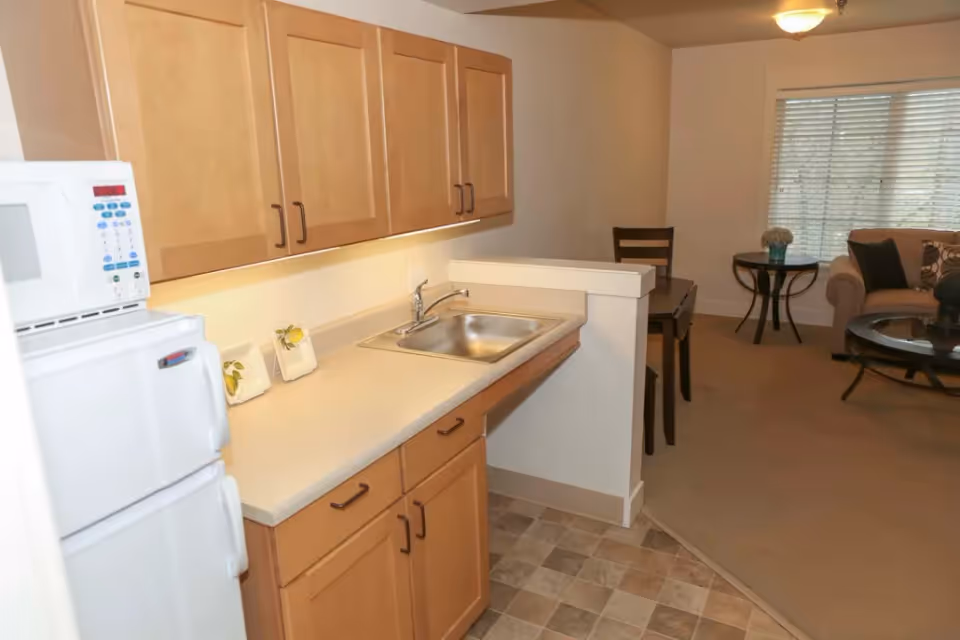 A small kitchen area with light wood cabinets, a white countertop, a stainless steel sink, a white microwave on top of a white refrigerator. Adjacent to the kitchen is a dining area with a dark wooden table and chairs, and a living room with a beige sofa, a round glass coffee table, and a side table with a vase of flowers near a window with blinds.