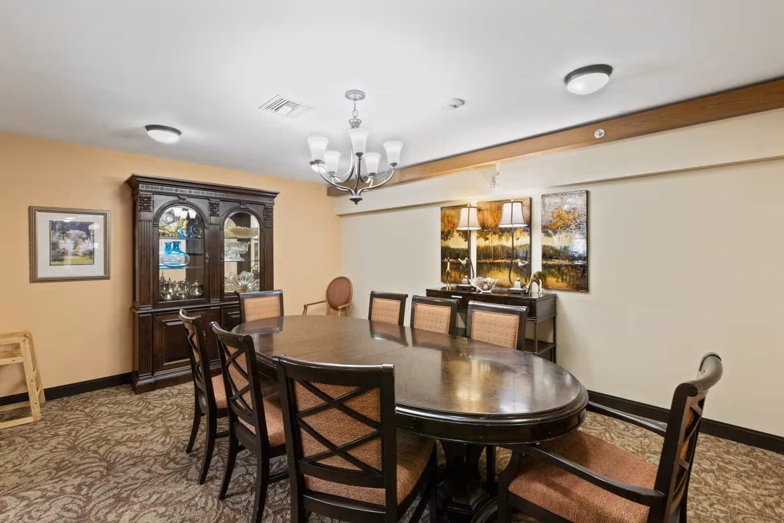 A formal dining room with a large oval wooden table surrounded by eight cushioned chairs. The room features a dark wooden china cabinet with glass doors displaying decorative items, a sideboard with two lamps and artwork above it, beige walls, and patterned carpet flooring. Ceiling lights and a chandelier provide illumination.