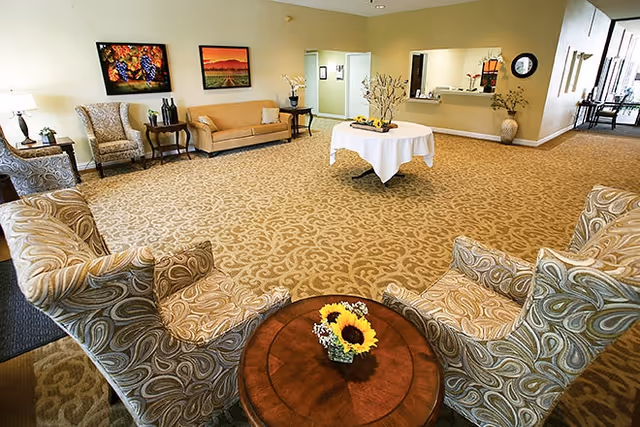A spacious and well-lit common area with patterned carpet, several upholstered chairs arranged around a round wooden table with a small sunflower centerpiece. In the background, there is a beige sofa, two armchairs, side tables with lamps, and framed artwork on the wall. A round table covered with a white cloth and decorative branches is placed in the center of the room. A reception window and hallway are visible in the back, with additional seating and natural light coming from the far end.