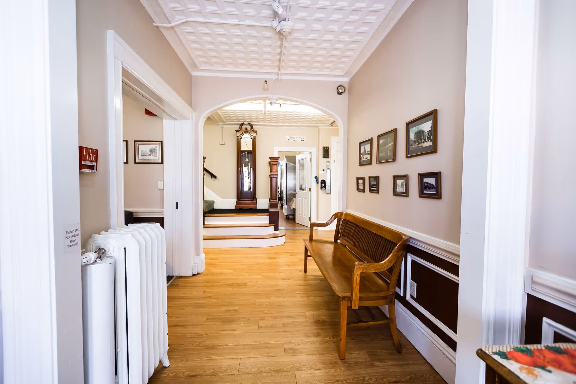 Interior hallway of Evergreen Place with wooden flooring, a wooden bench along the right wall, framed pictures hanging above the bench, a white radiator on the left, and a grandfather clock visible through an arched doorway at the end of the hallway.