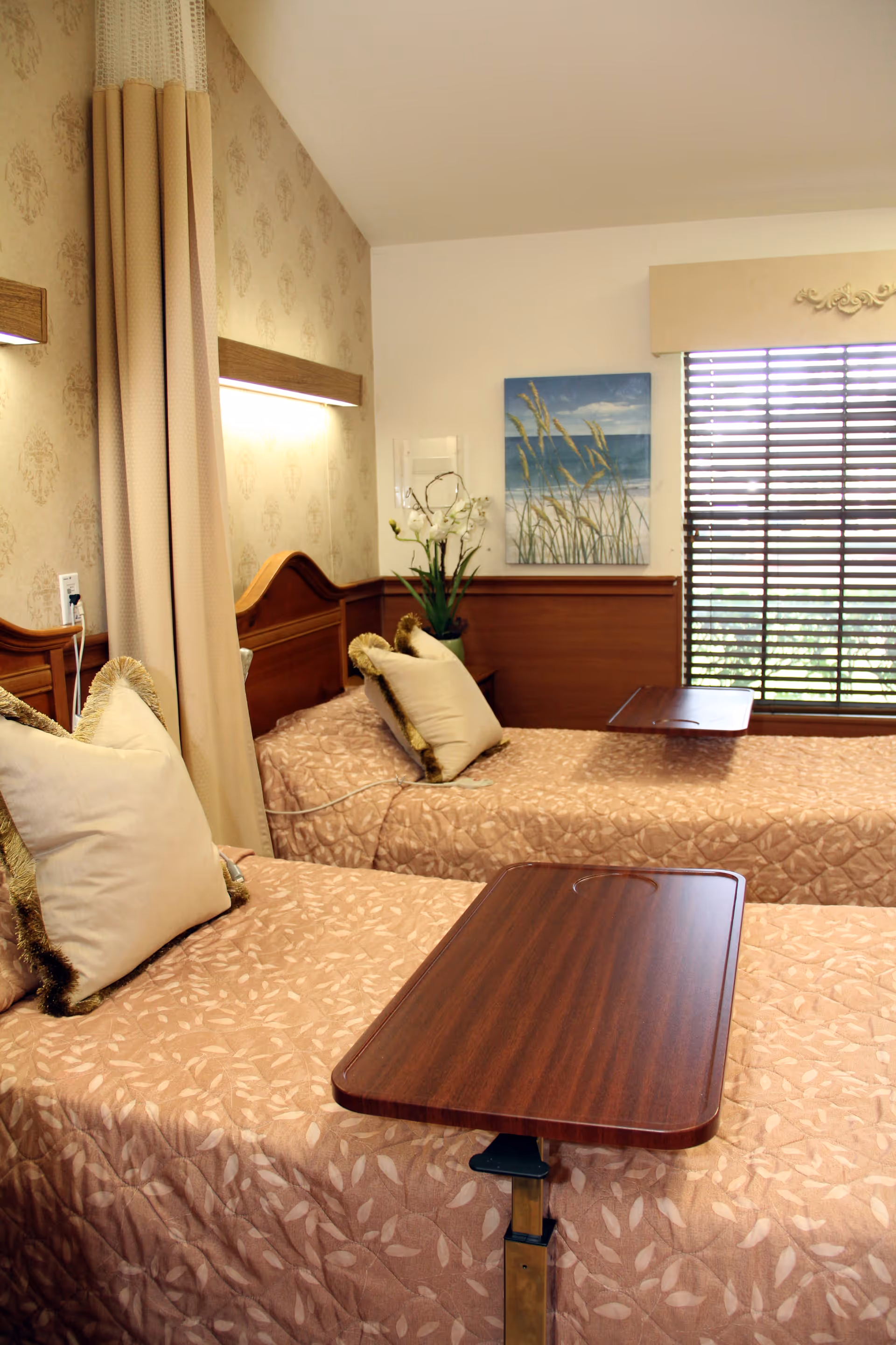 A well-lit room with two single beds covered in beige patterned bedspreads, each with a beige pillow with fringed edges. Each bed has a wooden overbed table positioned on top. The room features a window with horizontal blinds and a decorative valance, a painting of beach grass and ocean waves on the wall, and a vase with white flowers on a wooden ledge. The walls have a combination of beige patterned wallpaper and white paint.