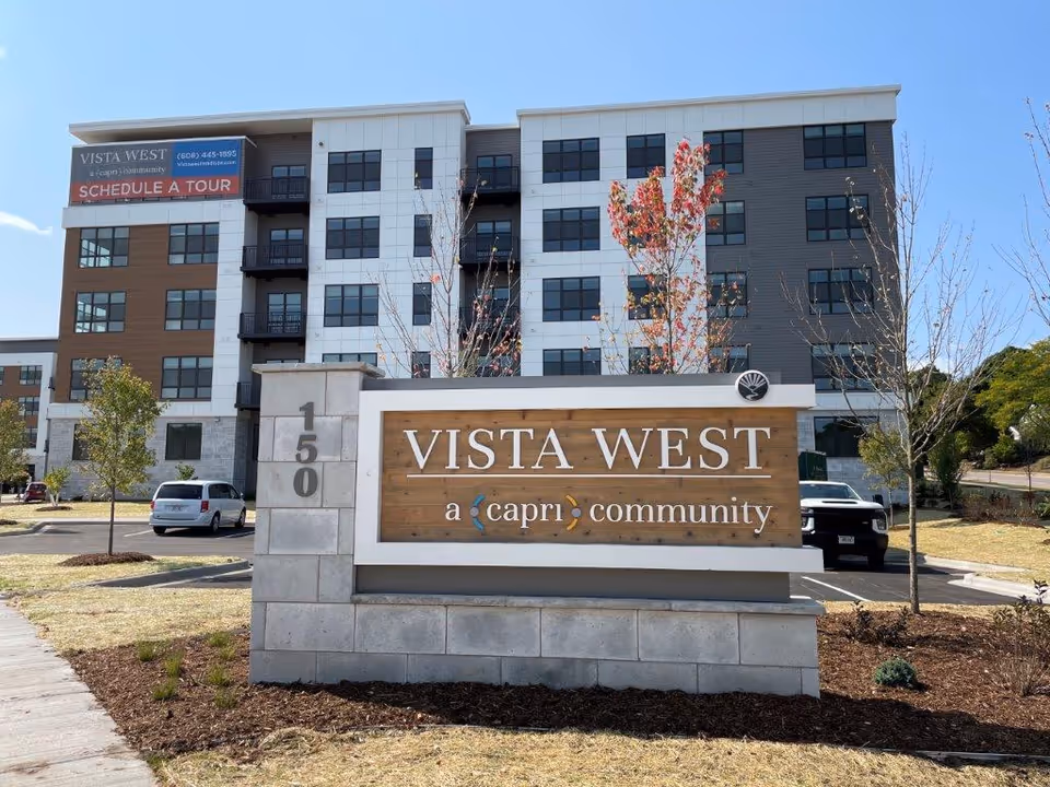 Exterior view of Vista West, a multi-story senior living community building with a large sign in front displaying the name 'Vista West a capri community' and the address number 150. The building has multiple windows and balconies, with some trees and parked cars visible around the property.