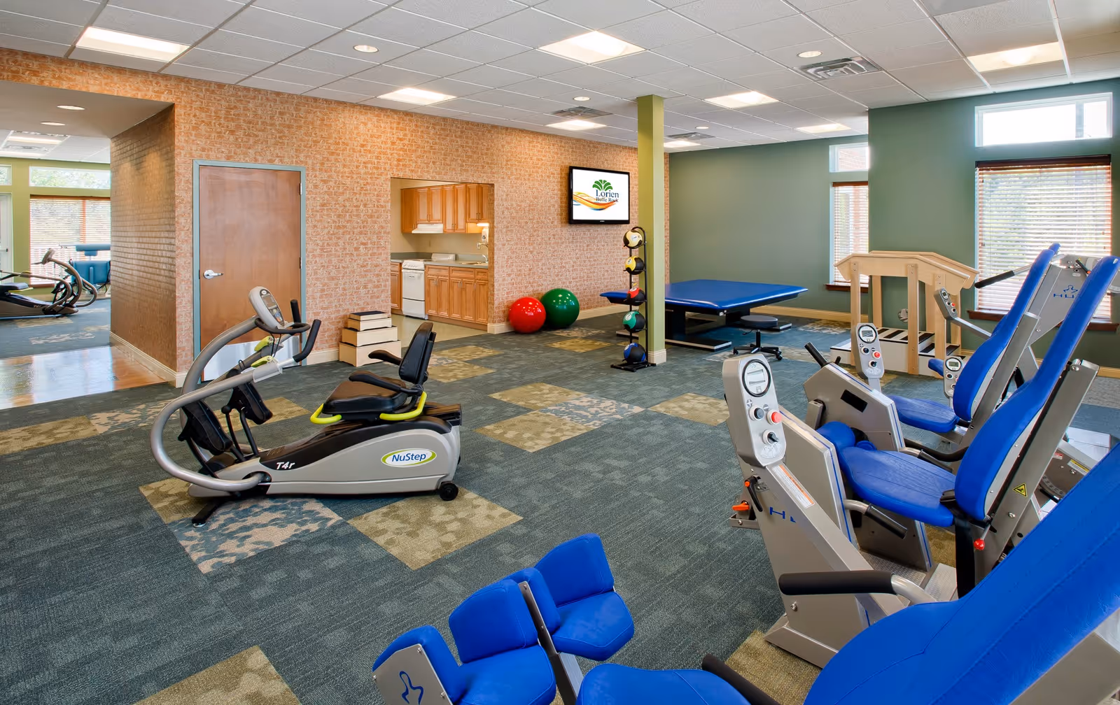 A spacious fitness room in a senior living facility featuring exercise equipment including NuStep machines, blue padded chairs, a therapy table, exercise balls, and a small kitchenette area in the background. The room has green and beige patterned carpet, green walls, and large windows with blinds allowing natural light.