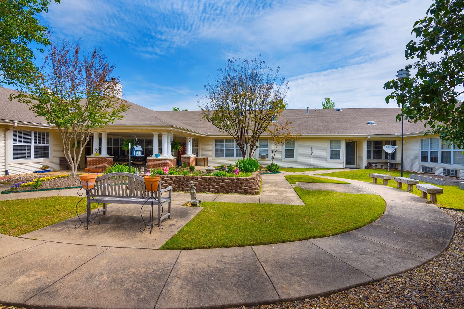 Outdoor courtyard area of a senior living facility with a curved concrete walkway, green grass, trees, flower beds, benches, and a building with multiple windows and a covered porch in the background under a partly cloudy blue sky.