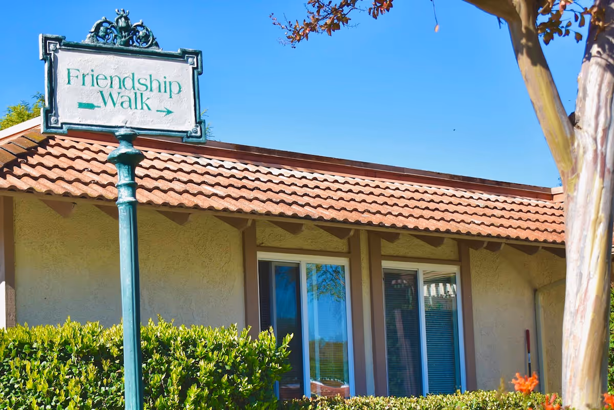 Outdoor scene at The Willows facility showing a green signpost with the text 'Friendship Walk' and arrows pointing left and right, in front of a building with a tiled roof, windows, and surrounded by bushes and a tree under a clear blue sky.