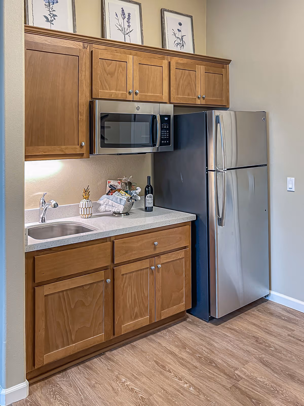 Small kitchen area with wooden cabinets, stainless steel refrigerator and microwave above a sink and countertop with decorative items.
