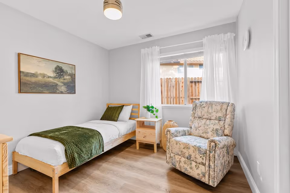A small, bright bedroom with a single wooden bed made with white linens and a green throw blanket. Next to the bed is a wooden nightstand with a small plant on it. A floral-patterned armchair is positioned near a window with sheer white curtains, and a landscape painting hangs on the wall above the bed. The floor is light wood, and the walls are painted white.